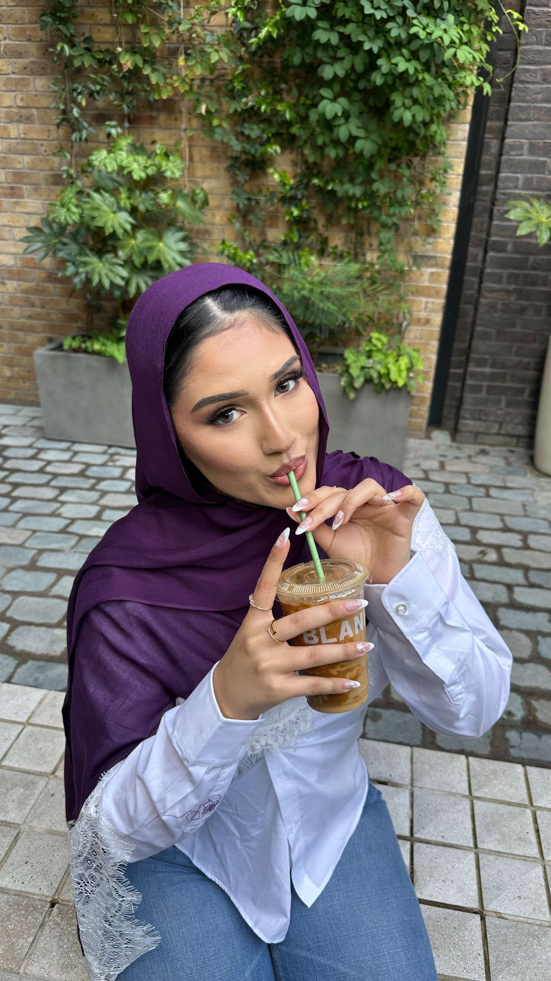 Portrait of a model in a purple hijab sipping iced coffee outside a café.