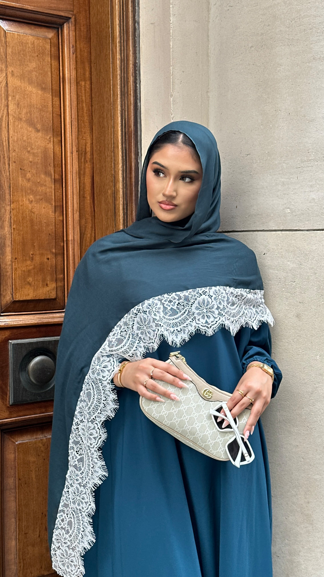 Aya the Label — model in a teal abaya and emerald lace-trim hijab holding a shoulder bag.
