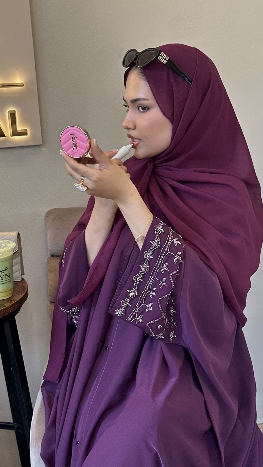 Model in a plum abaya applying lip product in a café, showing the beaded sleeve detail.